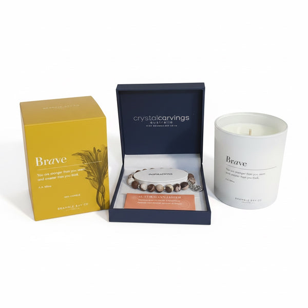 Crystal Carvings 'Brave' self-care gift set featuring a white candle, a yellow box, and a navy blue jewelry box containing a beaded bracelet and a rose gold card.