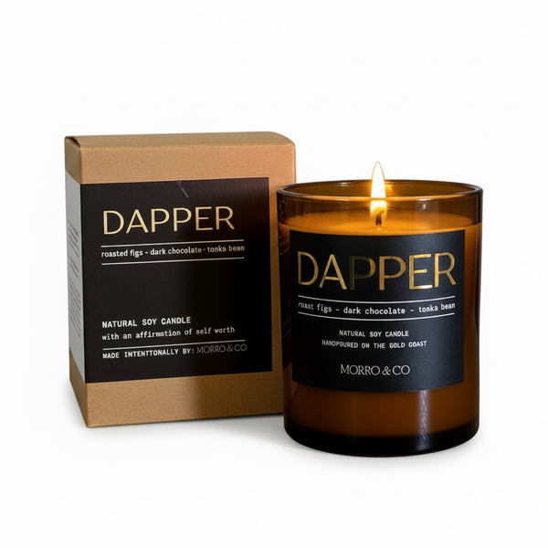 A lit amber glass jar candle with a black label reading DAPPER and MORRO & CO, shown next to its kraft paper box on a white background.