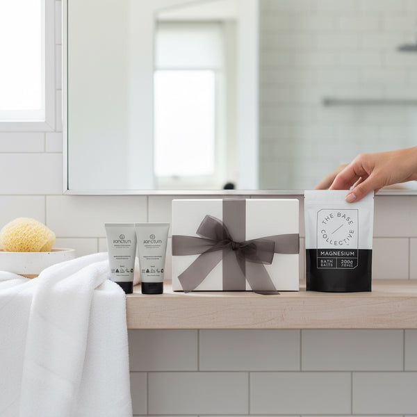 A bathroom gift set featuring a white box tied with a gray ribbon, two gray tubes from The Bath Collection, and a hand holding a packet of The Bath Collection Magnesium Bath Salt, set against a clean white tiled background.