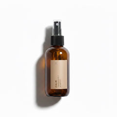 An amber glass spray bottle with a black pump dispenser and a beige label reading 'CALM' on a white background with a soft shadow.