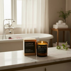 A lit DAPPER candle in a dark amber glass jar sits on a white marble bathroom counter next to its kraft paper box, with a softly blurred bathtub and warm-toned bathroom interior in the background.