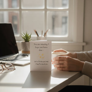 A white greeting card with black text and small gold stars stands on a desk in a bright room, next to a person's hands holding a white mug. A laptop, glasses, and small plant are visible in the background near a window.