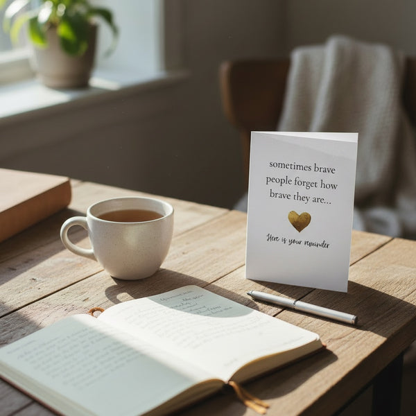 A white greeting card with black text and a small gold heart stands on a wooden table beside an open book, a white mug of tea, and a pen. The card reads, 'sometimes brave people forget how brave they are.