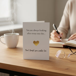 A white greeting card with black text and a gold heart stands on a wooden table in a cozy indoor setting. A person is writing in a notebook in the background, with a white mug, eyeglasses, and a pen visible.