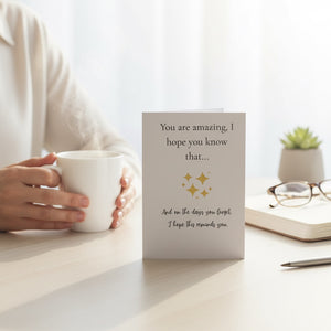 A vertical greeting card with a warm off-white background stands on a light wooden desk. The card features black cursive text that reads, 'You are amazing, I hope you know that.