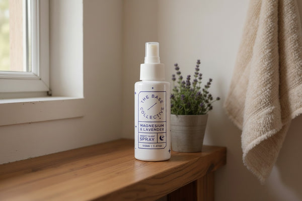 A white spray bottle labeled 'The Bath Collective' sits on a wooden windowsill next to a small potted lavender plant and a beige towel hanging on the wall.