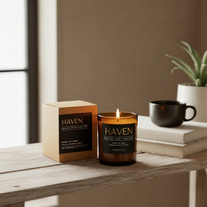 A lit amber glass candle labeled HAVEN sits on a wooden table next to its matching kraft paper box, with a black mug and books in the background.