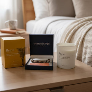 A cozy bedroom scene showing a candle product set on a wooden bedside table. The set includes a yellow Crystal Carvings 'Brave' box, an open navy blue Crystal Carvings gift box with a beaded bracelet inside, and a white glass jar candle labeled 'Brave' in a matching bedside setting with a bed and blanket 