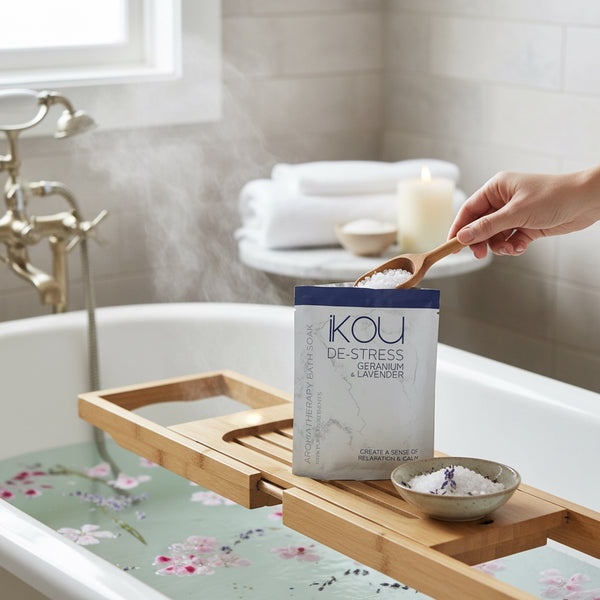 A hand uses a wooden spoon to scoop bath salts from a white and blue packet labeled 'ikou DE-STRESS GERMANY & LAVENDER' placed on a wooden bath caddy over a floral bathtub. Steam rises from the tub, with a candle and folded towels in the background.