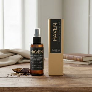 An amber glass spray bottle and its tall kraft paper box from MORRO & CO. HAVEN, placed on a wooden table with a neutral fabric and books in the background.