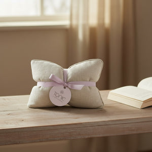 A beige fabric cushion-shaped product tied with a pale pink ribbon and a round tag reading 'TONIC', placed on a wooden table with an open book and soft window light in the background.