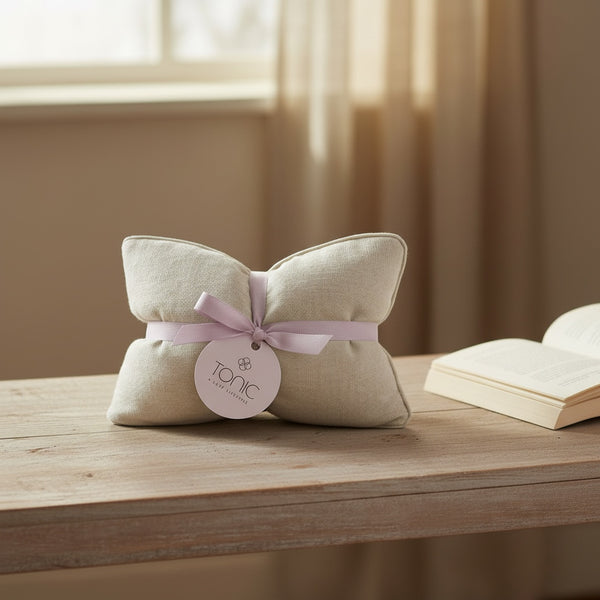 A beige fabric cushion-shaped product tied with a pale pink ribbon and a round tag reading 'TONIC', placed on a wooden table with an open book and soft window light in the background.