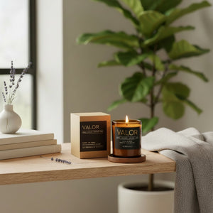 A lit VALOR soy candle in a dark amber glass jar sits on a wooden table next to its kraft paper box, styled in a cozy room setting with a potted fiddle-leaf fig plant and a gray throw blanket.