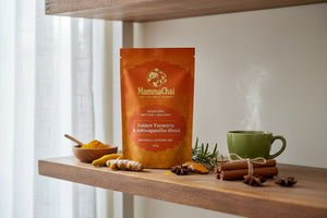 A stand-up pouch of MammaChai Golden Turmeric & Ashwagandha Blend herbal tea sits on a wooden shelf, styled with turmeric root, cinnamon sticks, star anise, rosemary, and a green mug with steam rising.