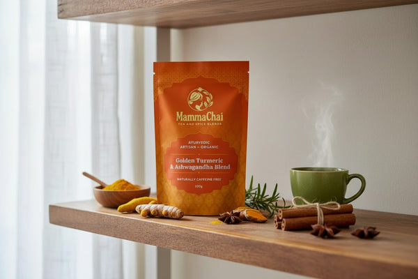 A stand-up pouch of MammaChai Golden Turmeric & Ashwagandha Blend herbal tea sits on a wooden shelf, styled with turmeric root, cinnamon sticks, star anise, rosemary, and a green mug with steam rising.