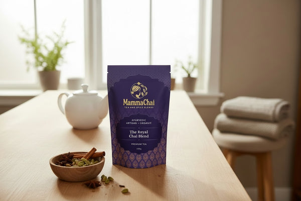 A purple stand-up pouch of MammaChai The Royal Chai Blend tea stands on a light wood table in a softly lit room, with a white teapot, small bowl of spices, and folded towels in the background.