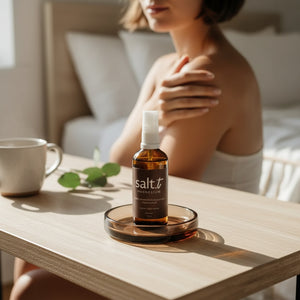 A small amber spray bottle labeled 'salt.t' sits on a round tray on a light wood table in a softly lit bedroom.