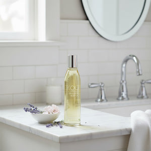 A bottle of KOU Haircare Moroccan Aloe Oil stands on a white marble bathroom counter next to a small bowl of lavender and pink flowers, with a white subway tile wall, chrome faucet, and round mirror in the background.