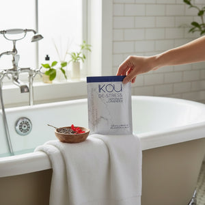 A hand holds a package of KOU De-Stress Lavender bath powder next to a white towel draped over the edge of a bathtub in a bright bathroom setting.