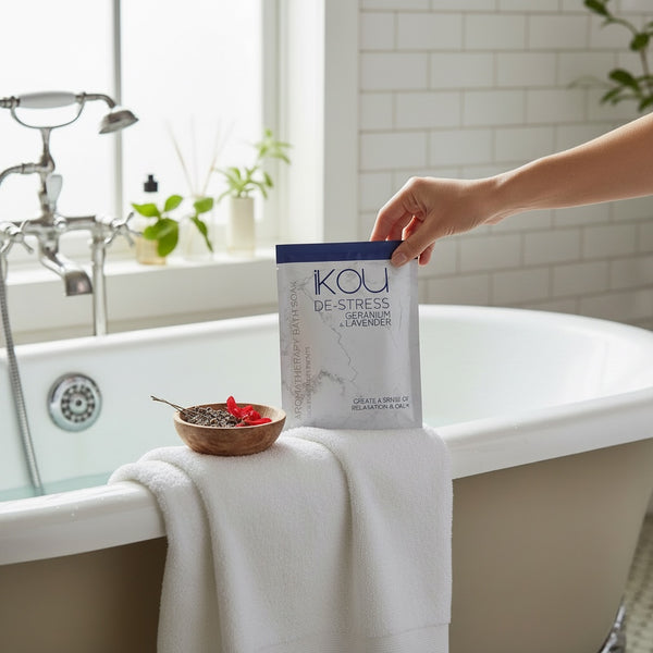 A hand holds a package of KOU De-Stress Lavender bath powder next to a white towel draped over the edge of a bathtub in a bright bathroom setting.