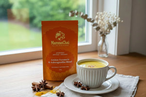 A stand-up pouch of MammaChai Golden Turmeric & Ashwagandha Blend tea stands on a wooden table beside a white cup of yellow tea on a saucer, with star anise and cinnamon sticks nearby and a blurred window background.