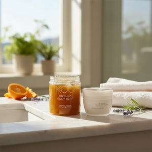 Two skincare products from the brand KOU are displayed on a white marble counter in a bright bathroom setting. The product on the left is a clear glass jar containing an orange-colored organic body buff with visible exfoliating granules, labeled 'KOU Organic Body Buff Orange & Citrus'.