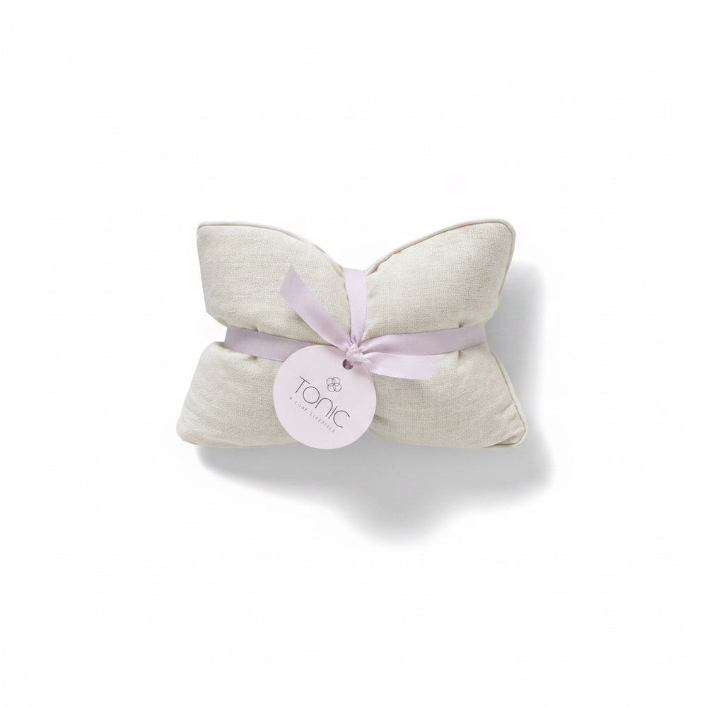 A folded cream-colored textured fabric pillow or cushion tied with a light lavender ribbon and a round white paper tag that reads 'tonic', set against a plain white background.