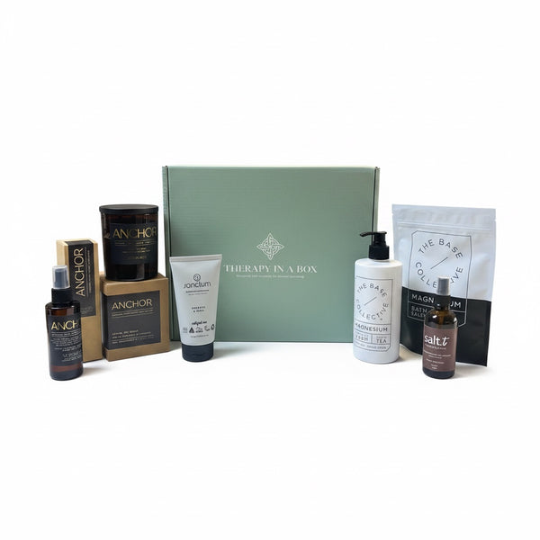 A curated wellness gift set arranged in front of a sage green box labeled 'Therapy in a Box.' The set includes amber bottles, white tubes, a black pump bottle, a white pouch, and a brown jar, with visible brands such as Auvik, Anchor, and The Bare Hand.