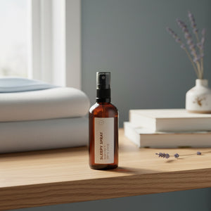 An amber glass spray bottle labeled 'SLEEPY SPRAY' stands on a light wood table in a softly styled bedroom setting with folded bedding, books, and lavender.