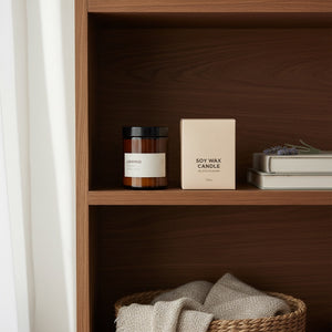 A soy wax candle in a cream-colored box sits on a wooden shelf next to an amber glass jar candle with a black lid and white label. The scene is styled with books and a woven basket below.