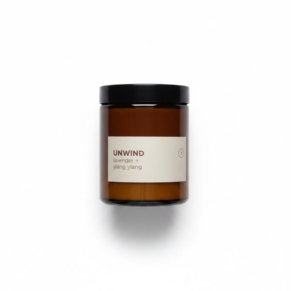 An amber glass jar candle with a black lid and a cream label reading 'UNWIND lavender + ylang ylang' on a white background.